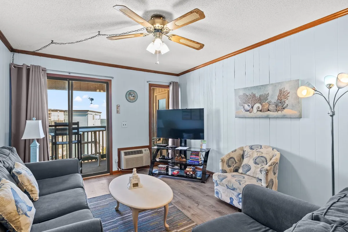 A comfortable living area featuring a plush sofa, coffee table, television, and stylish light fixtures. The space feels open and welcoming, with a suggestion of sunset views visible through an adjacent balcony door.