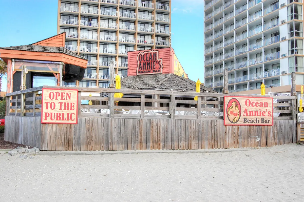 This image features the iconic Ocean Annie's Beach Bar sign and the beach bar's wooden structure, inviting guests to experience its laid-back, oceanfront vibe. The 'open to the public' sign highlights the bar's welcoming nature, and hints of the beach environment create a casual, lively atmosphere perfect for socializing, sipping cocktails, and enjoying live music.