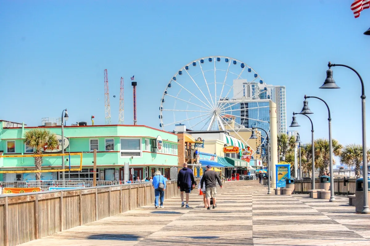 Colorful storefronts and amusements line the lively Myrtle Beach Boardwalk, with a towering Ferris wheel as a focal point. Palm trees, people enjoying rides, and vibrant signage create an atmosphere brimming with energy and family excitement, capturing the spirit of classic vacation fun.