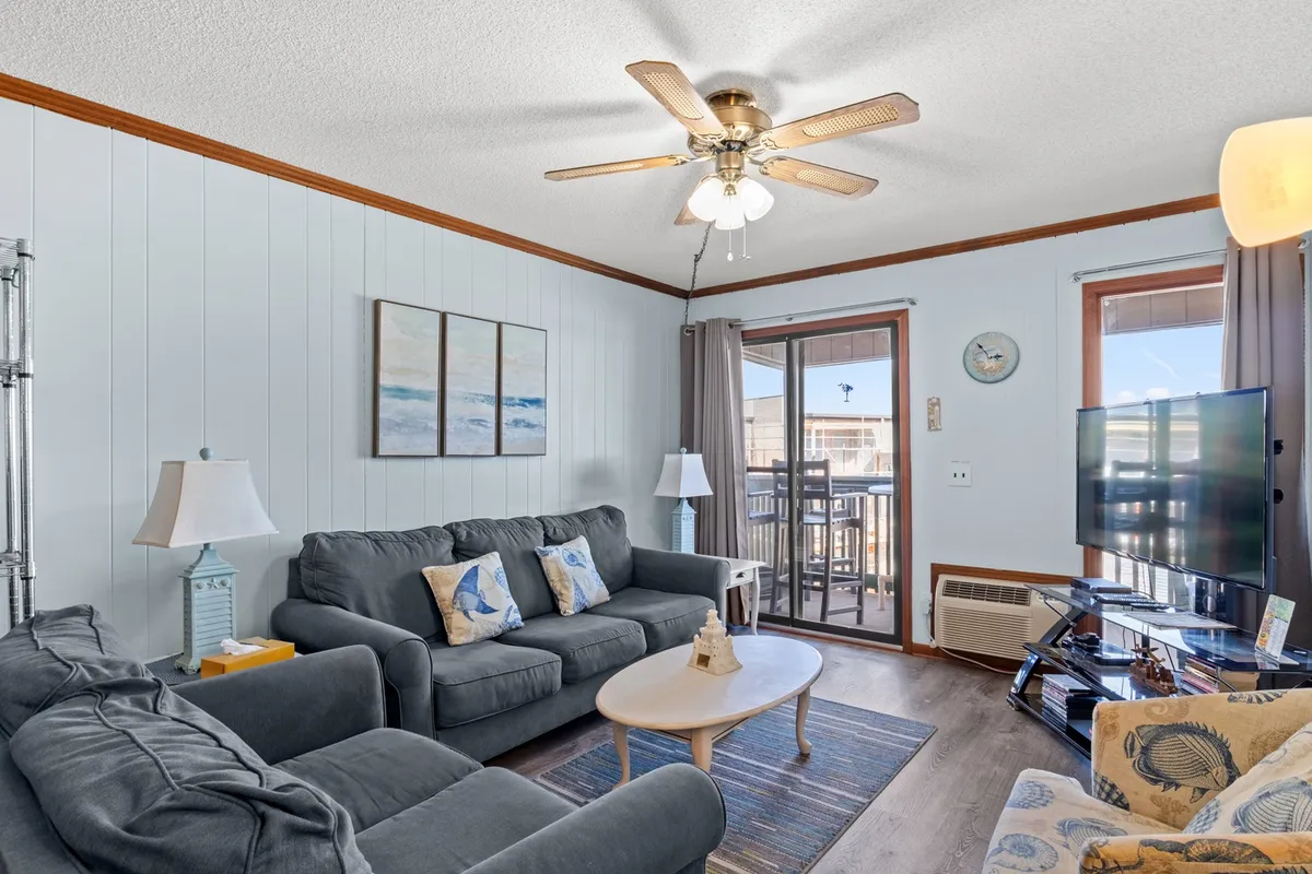 A cozy, spacious living room with comfortable sofas arranged around a coffee table, facing a large television. Artwork adorns the walls, and a prominent sliding glass door provides direct access to the balcony, allowing natural light to fill the room and offering a glimpse of the ocean beyond.