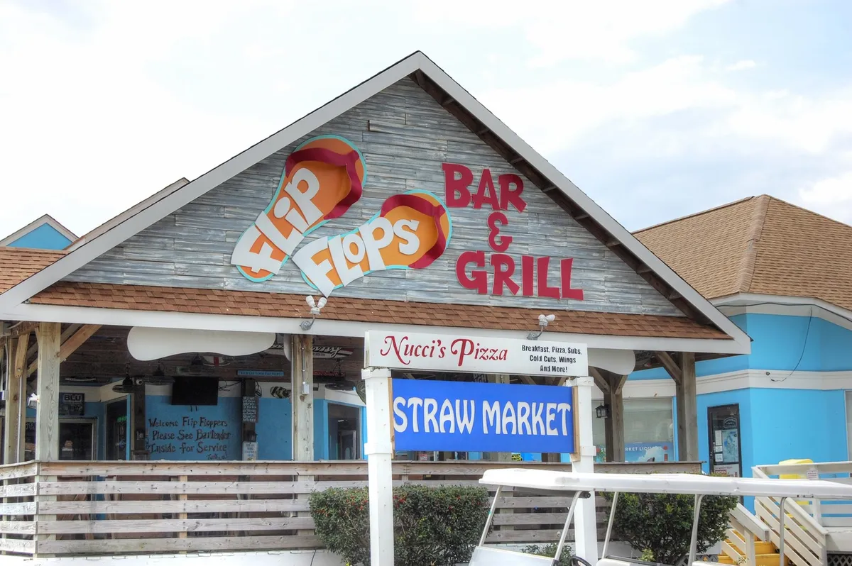 A lively exterior shot of multiple local establishments, including Flip Flops Bar & Grill, Nucci's Pizza, and the Straw Market. The vibrant signage and bustling atmosphere highlight the abundance of nearby options for dining and nightlife, further emphasizing the walkable and energetic character of Shore Drive.