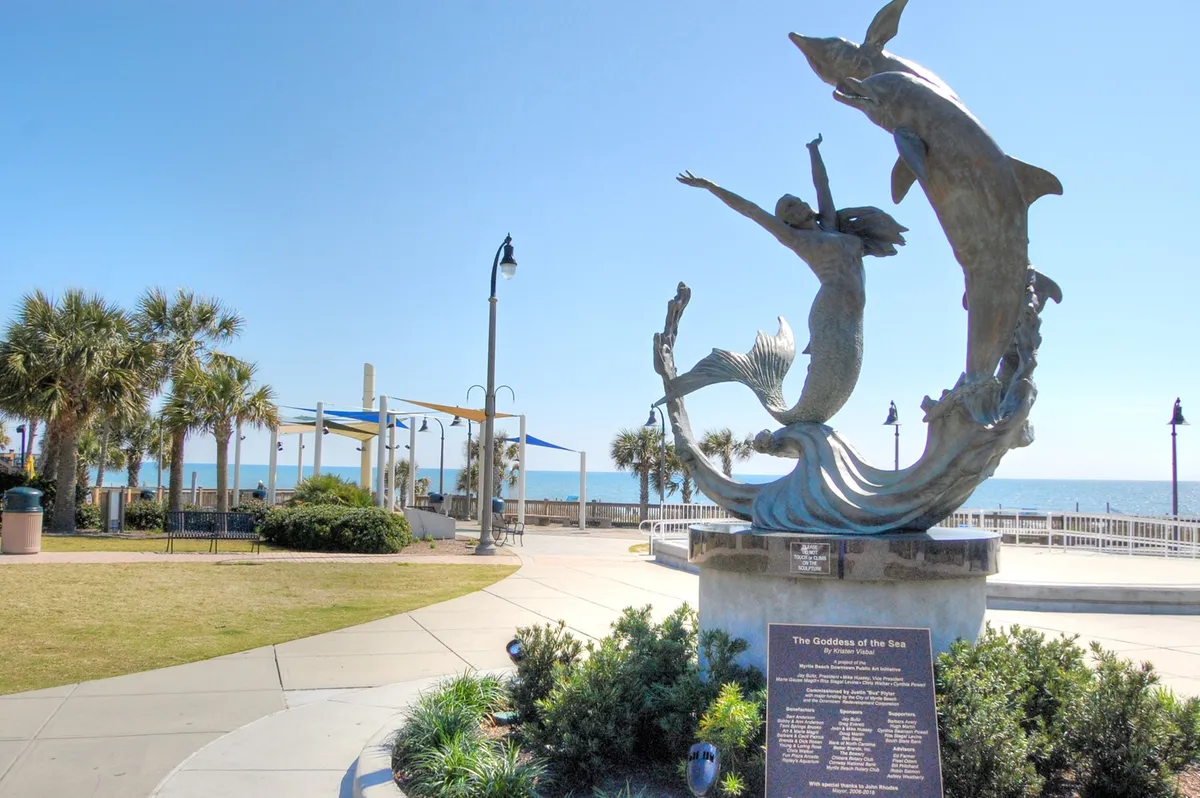 A picturesque oceanfront view featuring the Goddess of the Sea sculpture, palm trees, and a serene beachfront setting. Visitors can be seen walking, relaxing, and enjoying the natural beauty of the coast.