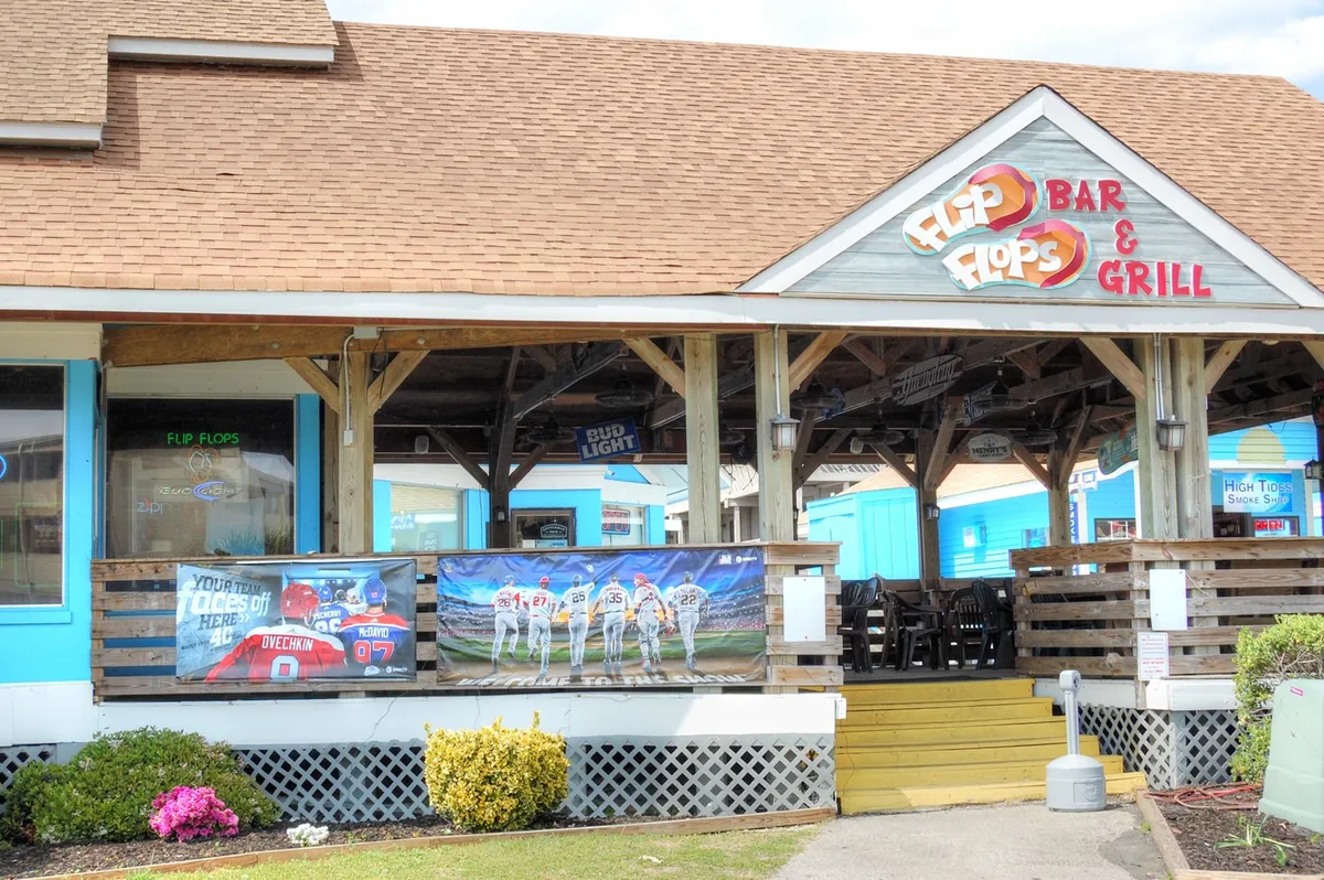 The exterior of Flip Flops Bar & Grill is captured here, featuring clear signage, promotional banners, and a spacious outdoor seating area. The scene suggests a lively, social environment perfect for group gatherings and casual nights out, with hints of local nightlife activity and the potential for events like live sports or themed nights.