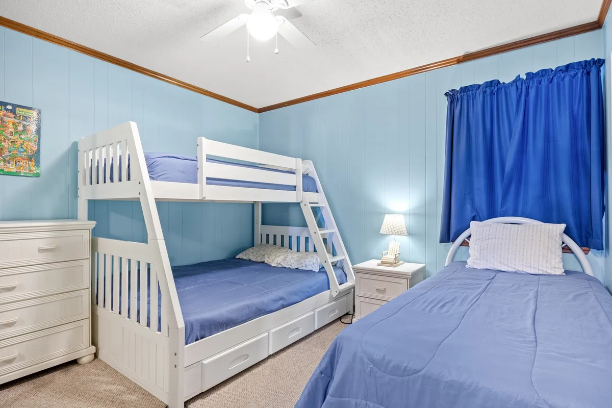 A playful and inviting bedroom outfitted with a bunk bed and a single bed, both with blue bedding. A nightstand with a lamp and a dresser offer ample storage, while blue curtains add a cheerful touch. The space is ideal for accommodating families or groups with children.