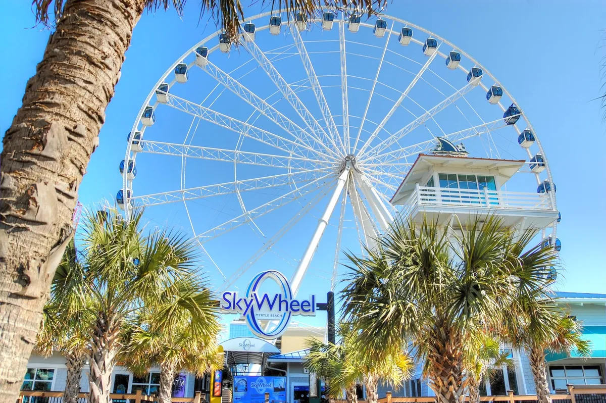 The iconic SkyWheel ferris wheel rises above palm trees and the entrance plaza, inviting guests to experience breathtaking panoramic views of the Myrtle Beach coastline. The area is bustling with visitors enjoying the vibrant local atmosphere.