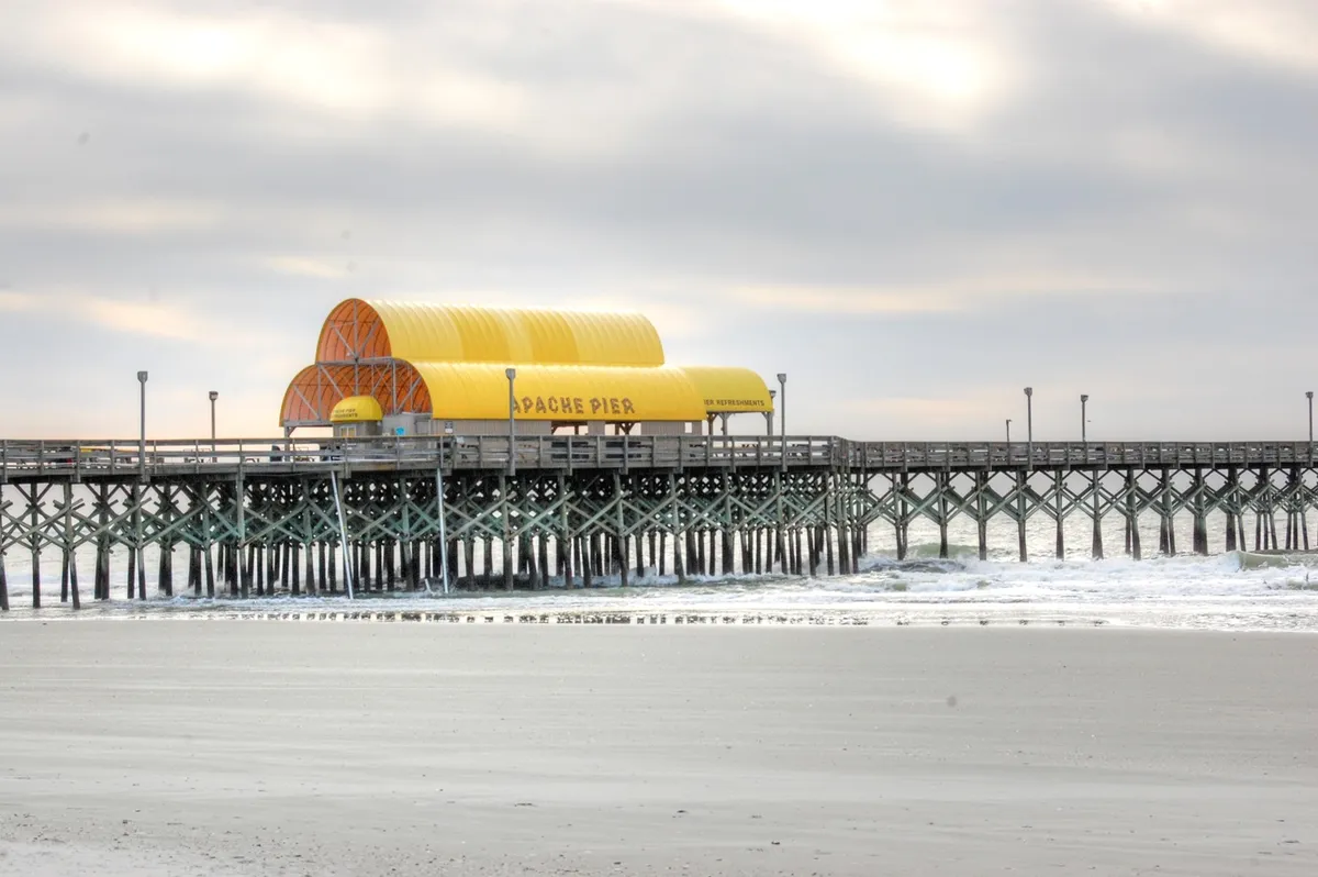 The Apache Pier stretches out over the Atlantic, its yellow-roofed pavilion standing above wooden planks. Visitors stroll, fish, and photograph the ocean, all while bathed in the serene light of sunrise or sunset, capturing the leisurely and scenic side of Myrtle Beach life.